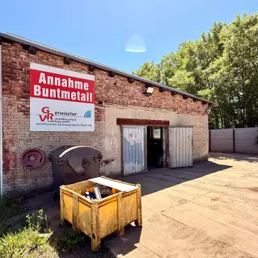 Ein Gebäude mit dem Schild „Annahme Buntmetall“ und einem gelben Sammelcontainer vor dem Eingang.