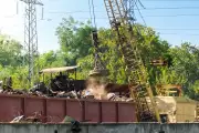 Ein Kran hebt Metallschrott aus einem Container auf einem Recyclinghof. Im Hintergrund sind Grünflächen und Stromleitungen zu sehen.